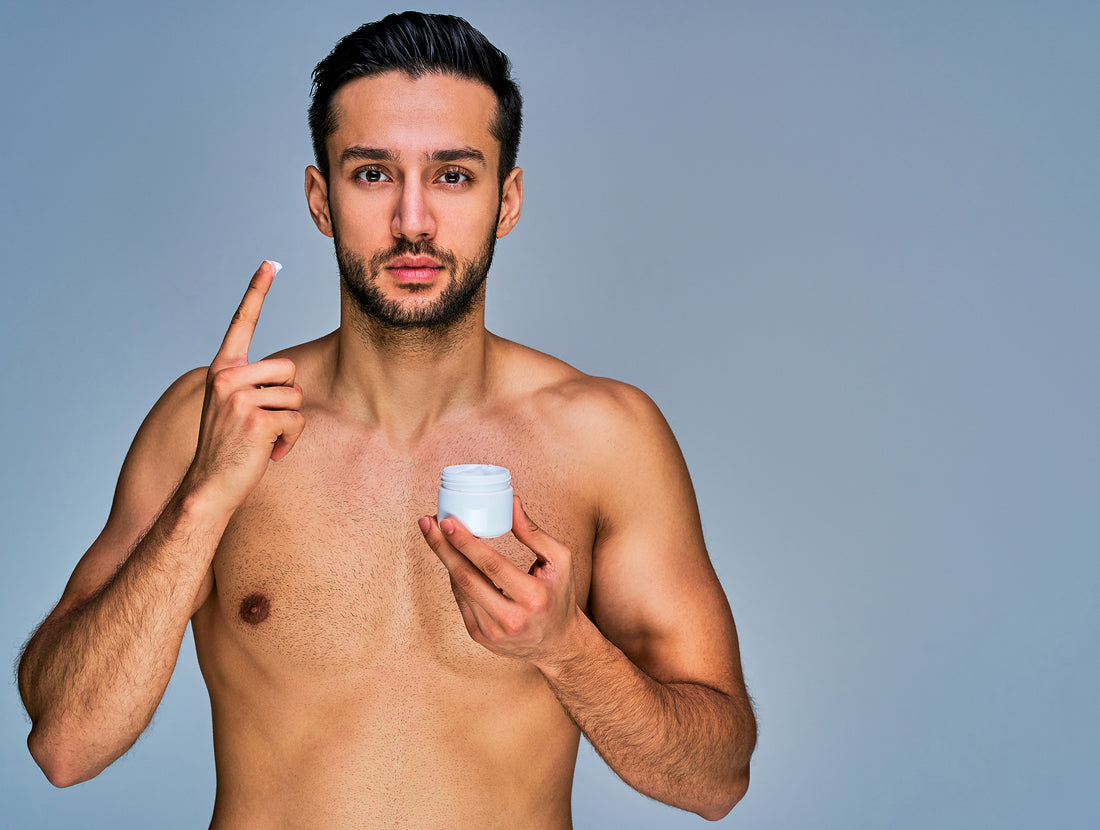 A young, shirtless, bearded man about to apply a cream to his face. Photo Credit: IC Production / Getty Images