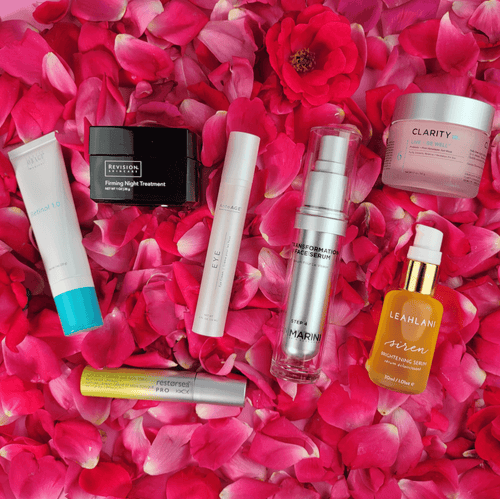 Several skincare products laying on a bed of dark pink rose petals.