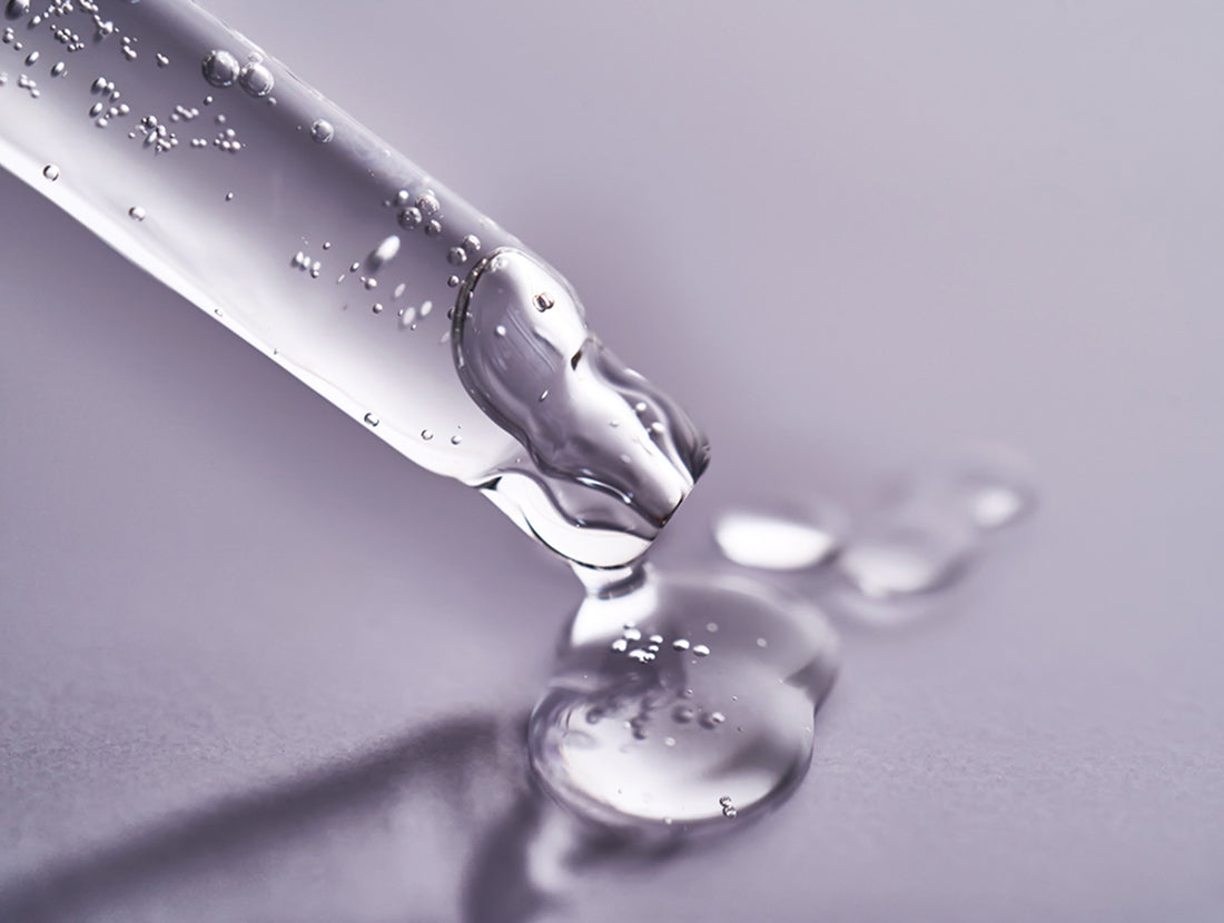 Closeup on a glass eyedropper releasing a drop of clear serum onto a grey-purple background. Photo Credit: Mariya Borisova / iStock Photo