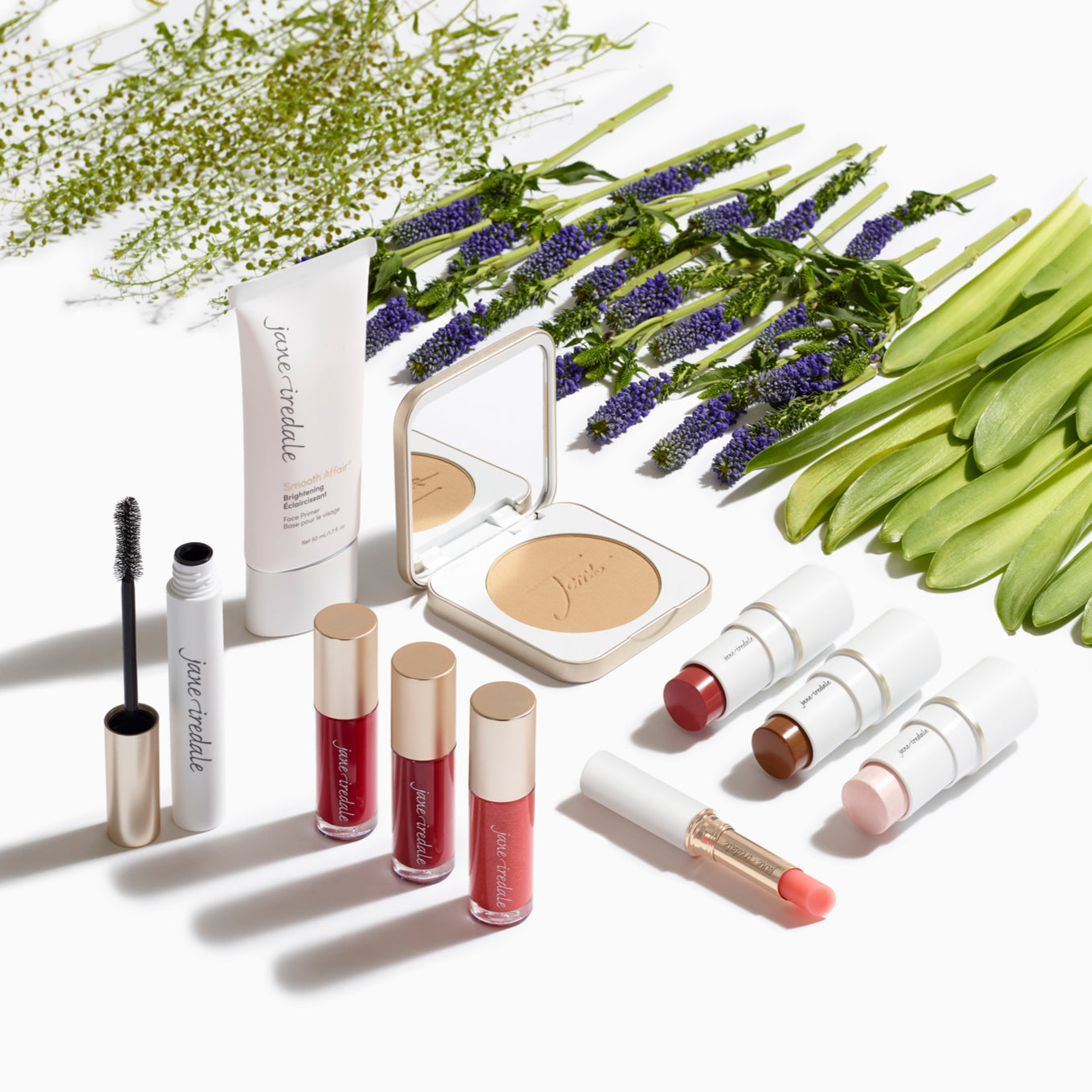 Several jane iredale products artistically arranged on a white background with some flowers and leaves lined up nearby