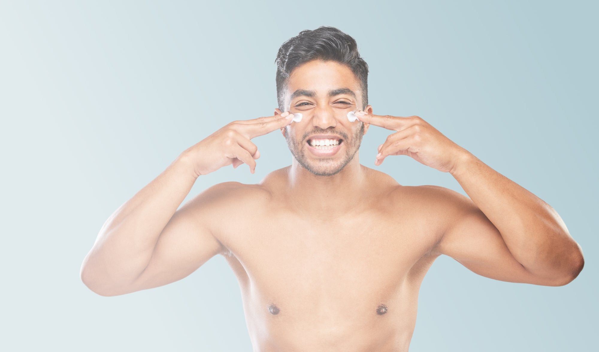 A young, shirtless man smiles as he applies a white cream under his eyes