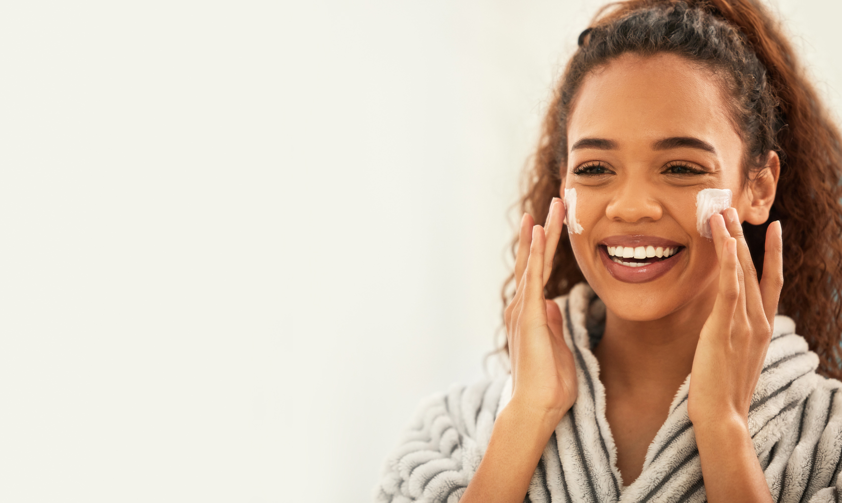 A young woman wearing a striped bathrobe smiles as she applies a white cream to her face.