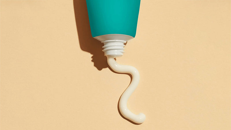 A turquoise colored tube squeezing out a white cream that contains Lactic acid on a tan background